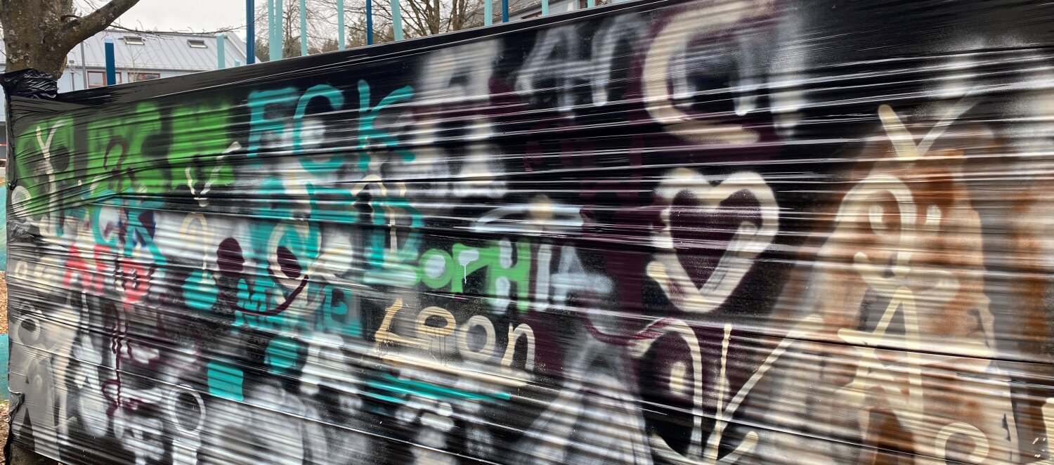 Hier ist eine schwarze Plane zu sehen, die zwischen einen Baum und eine Laterne gespannt wurde. Auf dieser Plane sind verschiedene Graffitis zu sehen. 