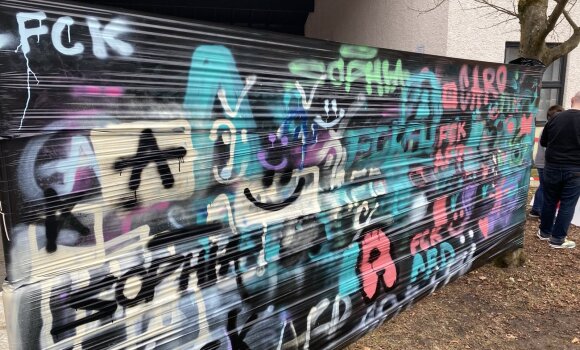 Hier ist eine schwarze Plane zu sehen, die zwischen einen Baum und eine Laterne gespannt wurde. Auf dieser Plane sind verschiedene Graffitis zu sehen. 