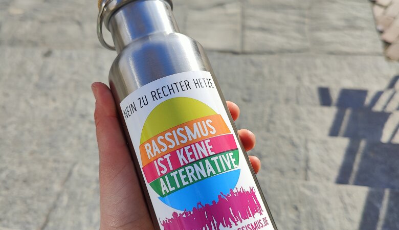 Zu sehen ist eine silberne Isolierkanne mit einem Sticker auf dem steht "Rassismus ist keine Alternative".